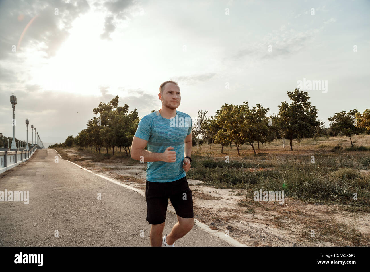 Portrait of Caucasian guy dans un tee-shirt bleu et un short noir qui s'entraîne et s'exécute sur la piste asphaltée pendant le coucher du soleil Banque D'Images