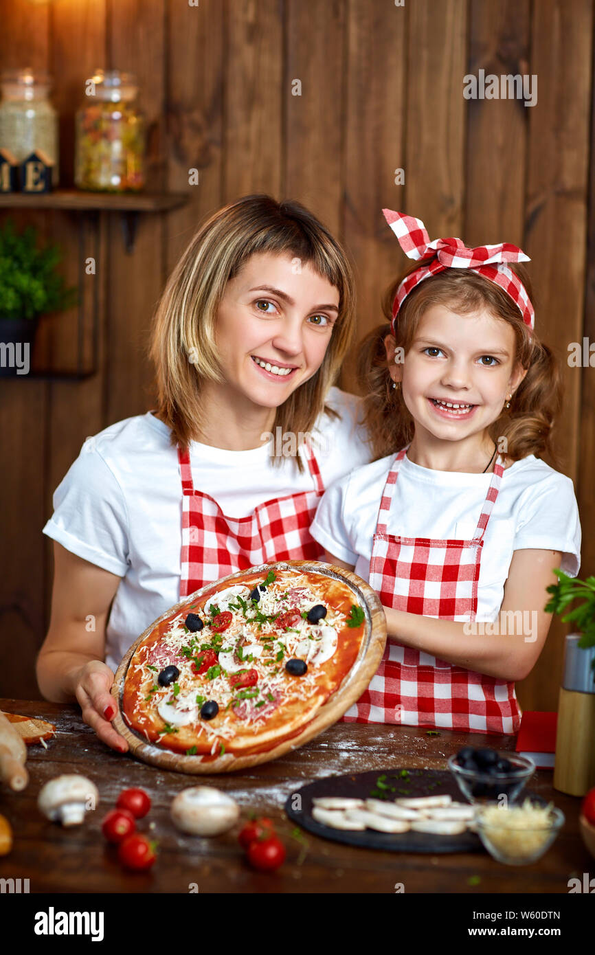 Happy happy mother and daughter wearing white T-Shirts et tabliers à carreaux montrant pizza cuite et looking at camera dans la belle cuisine en bois Banque D'Images