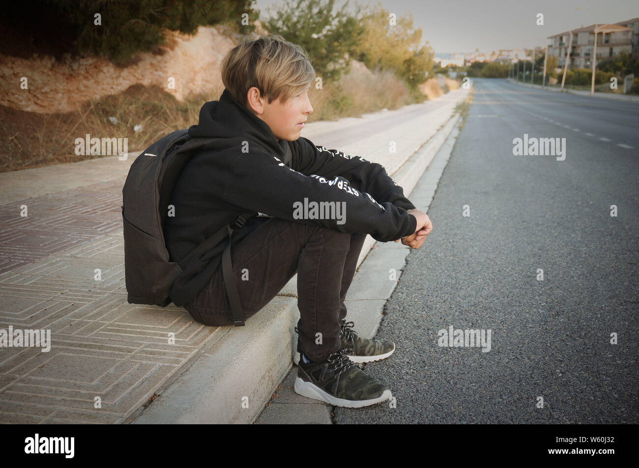 Blonde teenage boy sur route asphaltée vêtu de noir vêtir et sac à dos Banque D'Images