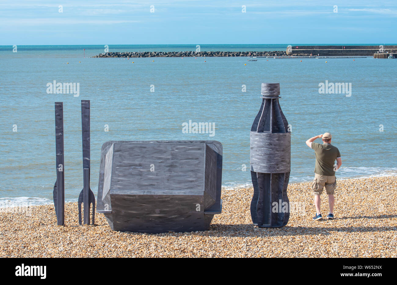 Lyme Regis, dans le Dorset, UK. Le 31 juillet 2019. Météo France : un homme admire la vue sur une litière géante sculpture d'emballage à emporter qui a été érigée sur la pittoresque plage de la station balnéaire de Lyme Regis. Composé d'un plastique d'une hauteur de 8 pieds, couverts de bouteilles de boissons mesurant 6 pieds de haut et une puce/burger fort de 4x4 pied les sculptures de promouvoir les efforts de la litière de côte et mer et partenaires à la promotion de choix écologiques. Il est à espérer que l'installation temporaire par Raisey Jack concepteur architectural va encourager les gens à prendre leur litière maison. Credit : Banque D'Images