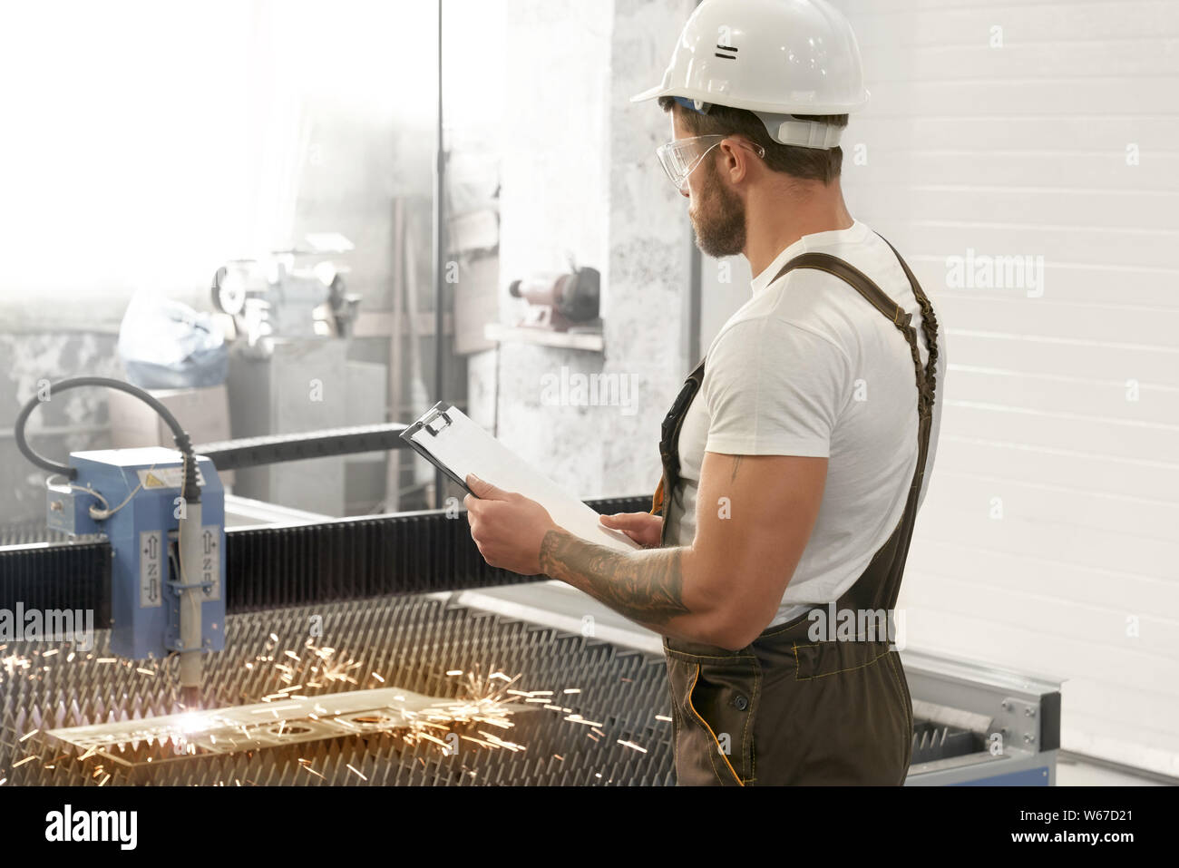 Voir du côté de l'ingénieur homme en uniforme, lunettes de protection et maintien du casque et dossier à la découpe plasma à. Processus de contrôle de l'homme musclé des dispositifs et équipements. Banque D'Images