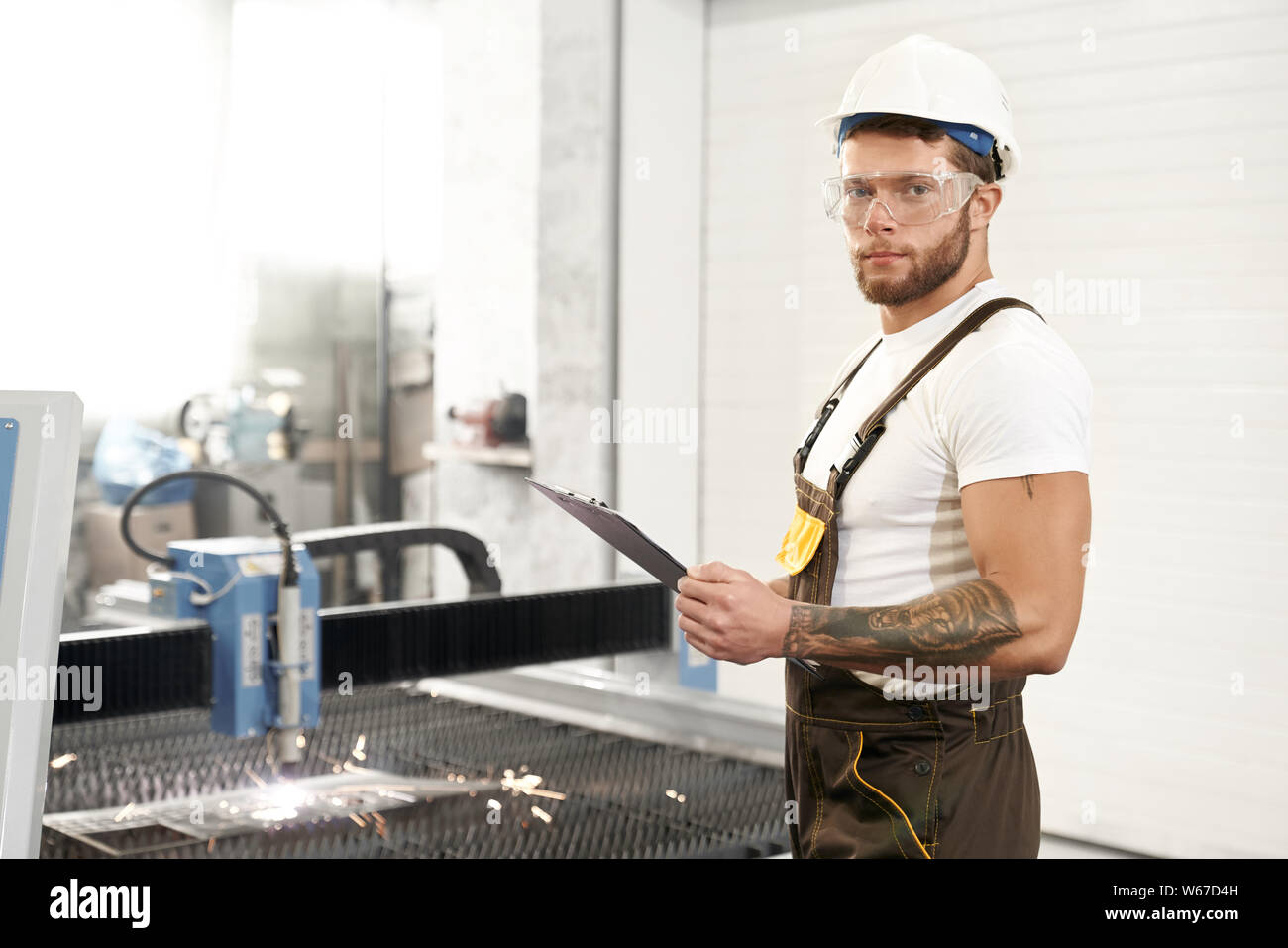 Vue latérale d'un mécanicien dans des lunettes de protection, casque et uniforme à la caméra et à poser tout en travaillant à l'usine de métal. Ingénieur mâle debout près de CNC et de coupage plasma de contrôle. Banque D'Images