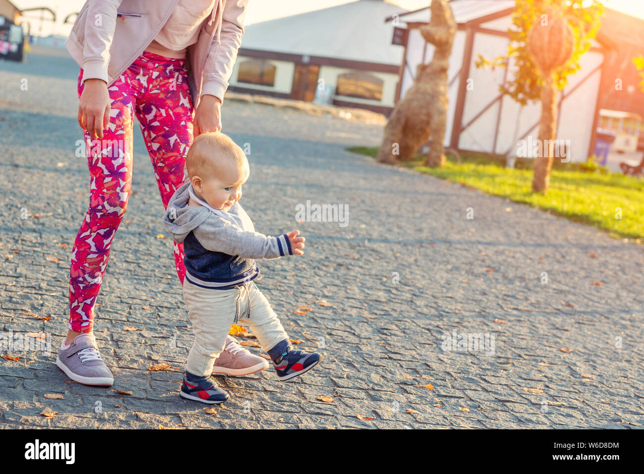 Jolie petite blonde adorable bébé garçon faire les premières étapes avec le soutien aux mères au parc de la ville à l'heure du coucher du soleil en soirée. Happy funny enfant l'apprentissage de la marche Banque D'Images