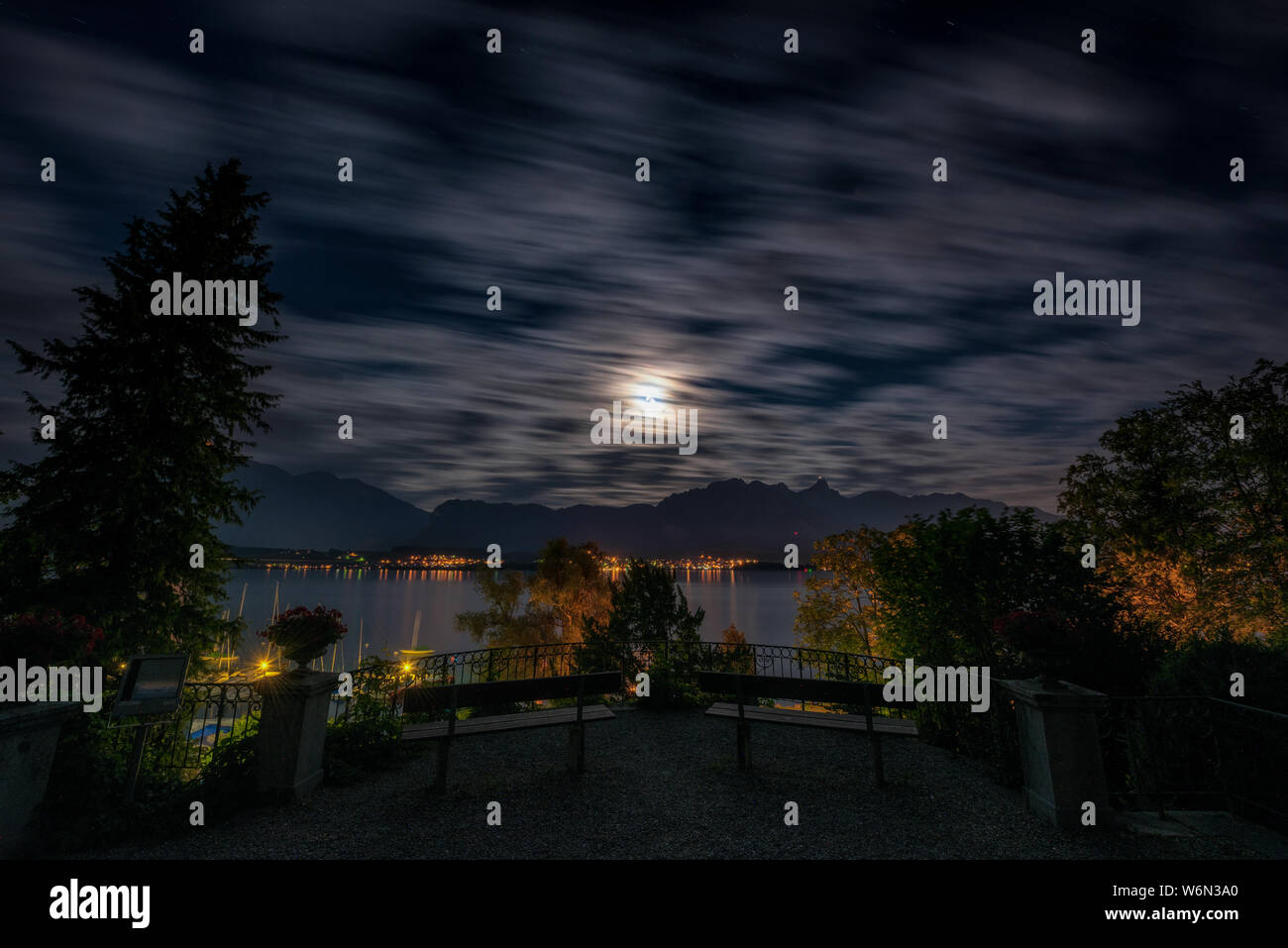 Une longue exposition d'un ciel nocturne avec franges de lune sur le Stockhorn de montagnes, le lac de Thoune, Hilterfingen marina et deux bancs et garde-corps de la parc du château Hünegg Banque D'Images