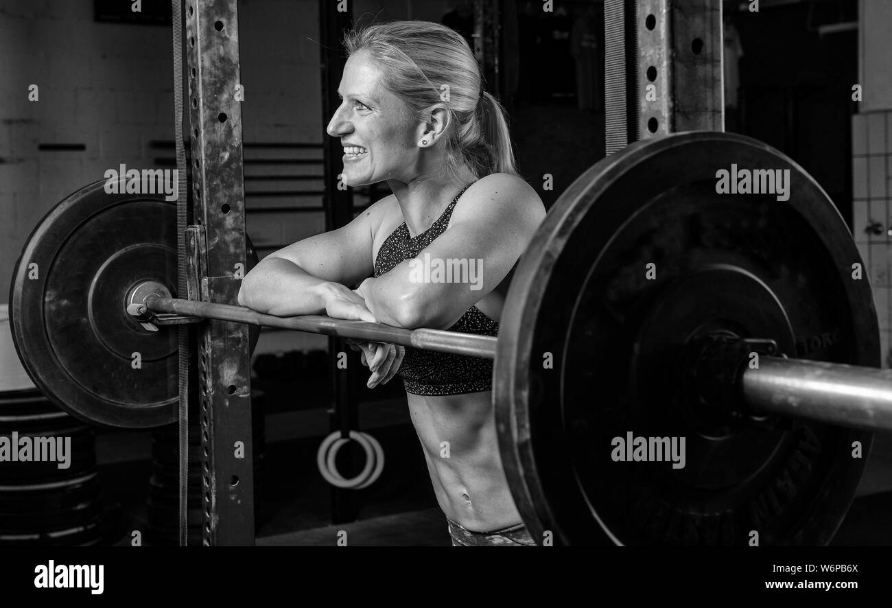 Portrait noir et blanc d'une belle femme avec de fortes abs. L'athletic et smiling blonde femme est appuyée contre le rack d'haltères longs. Banque D'Images