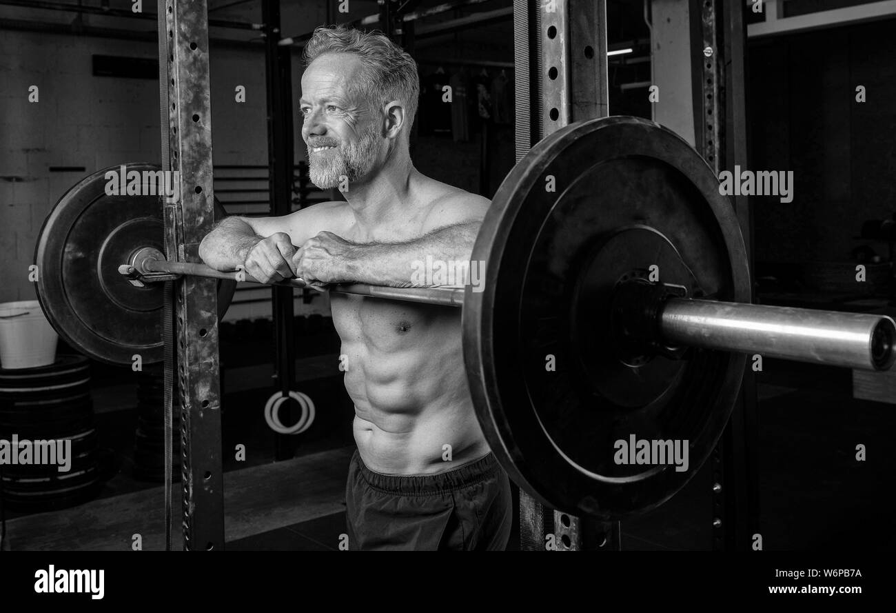 Le noir et blanc portrait d'un athlète d'âge moyen musculaire avec Sixpack. L'homme barbu souriant et athlétique est appuyé contre le rack d'haltères longs. Banque D'Images