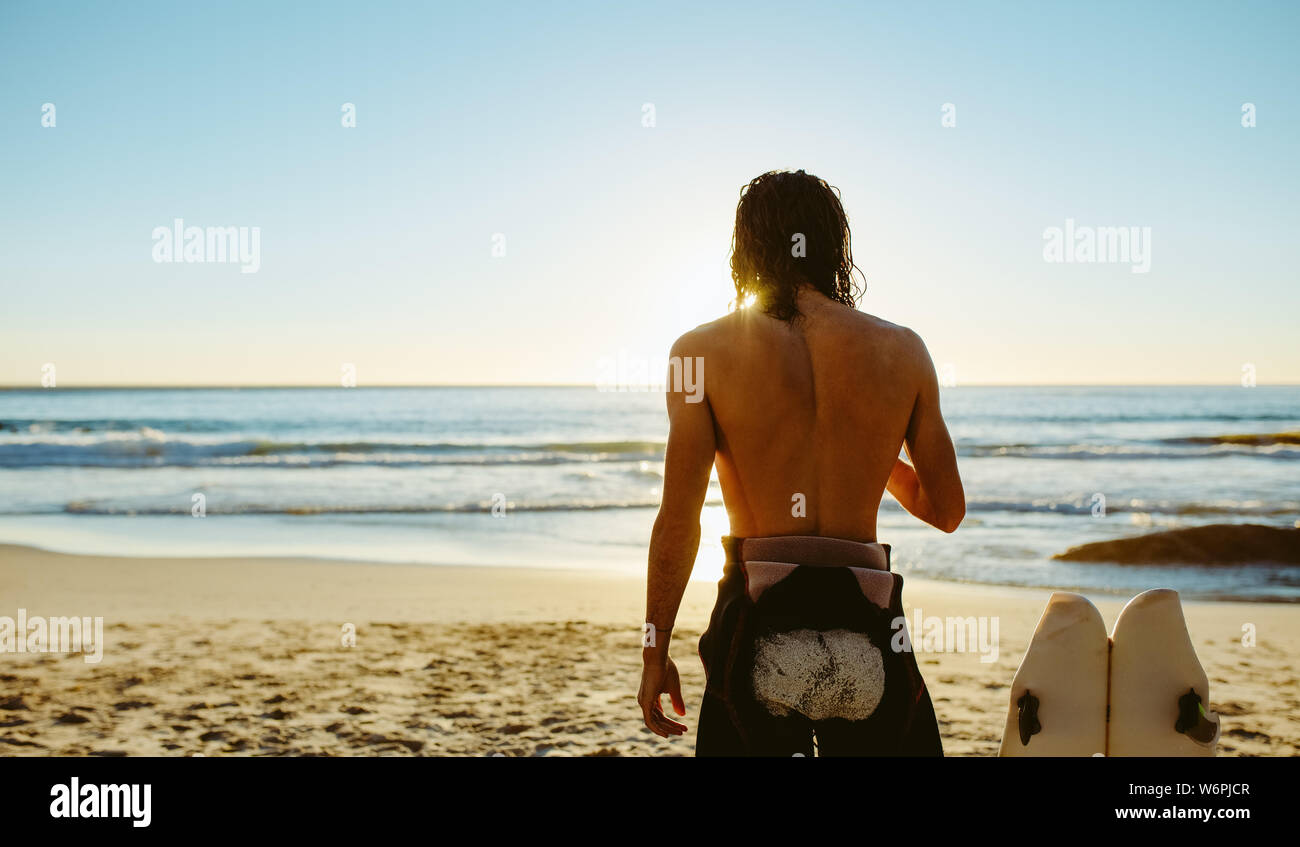 Vue arrière coup de jeune homme debout sur la plage avec planche de surf. Surfeur homme détente sur la mer un jour d'été. Banque D'Images