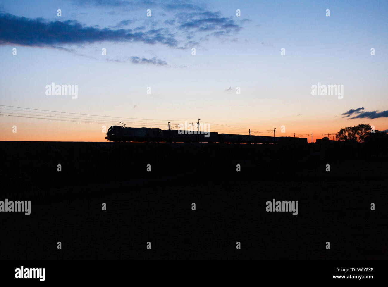 Services ferroviaires directs 88 locomotive électrique sur la ligne principale de la côte ouest de faire une silhouette au coucher du soleil avec le Stobart / train de marchandises Tesco Banque D'Images