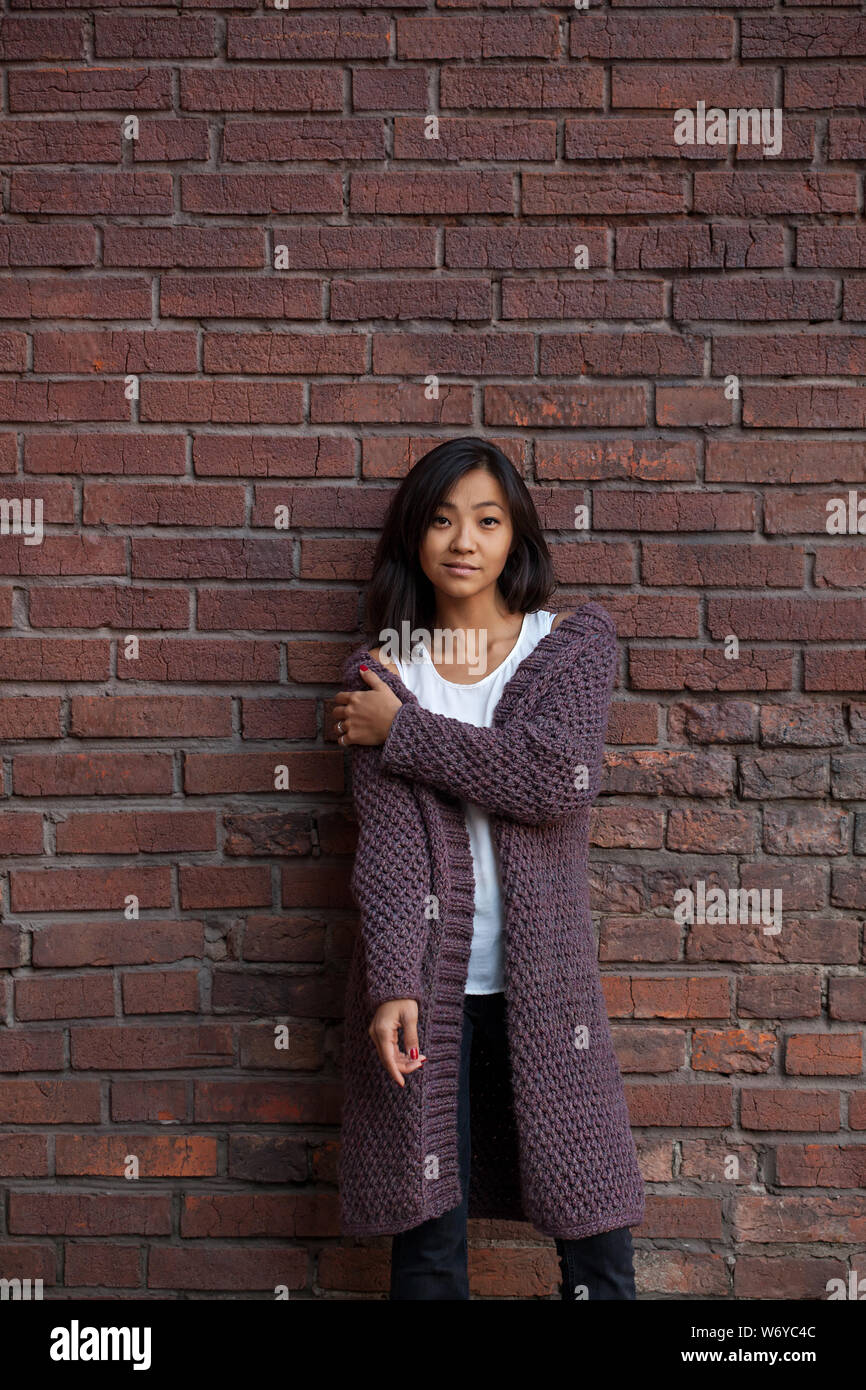 Belle asiatique Jeune femme debout dans un violet tricoté cardigan chaud, serrant elle-même sur l'épaule d'une main contre un mur permanent Banque D'Images