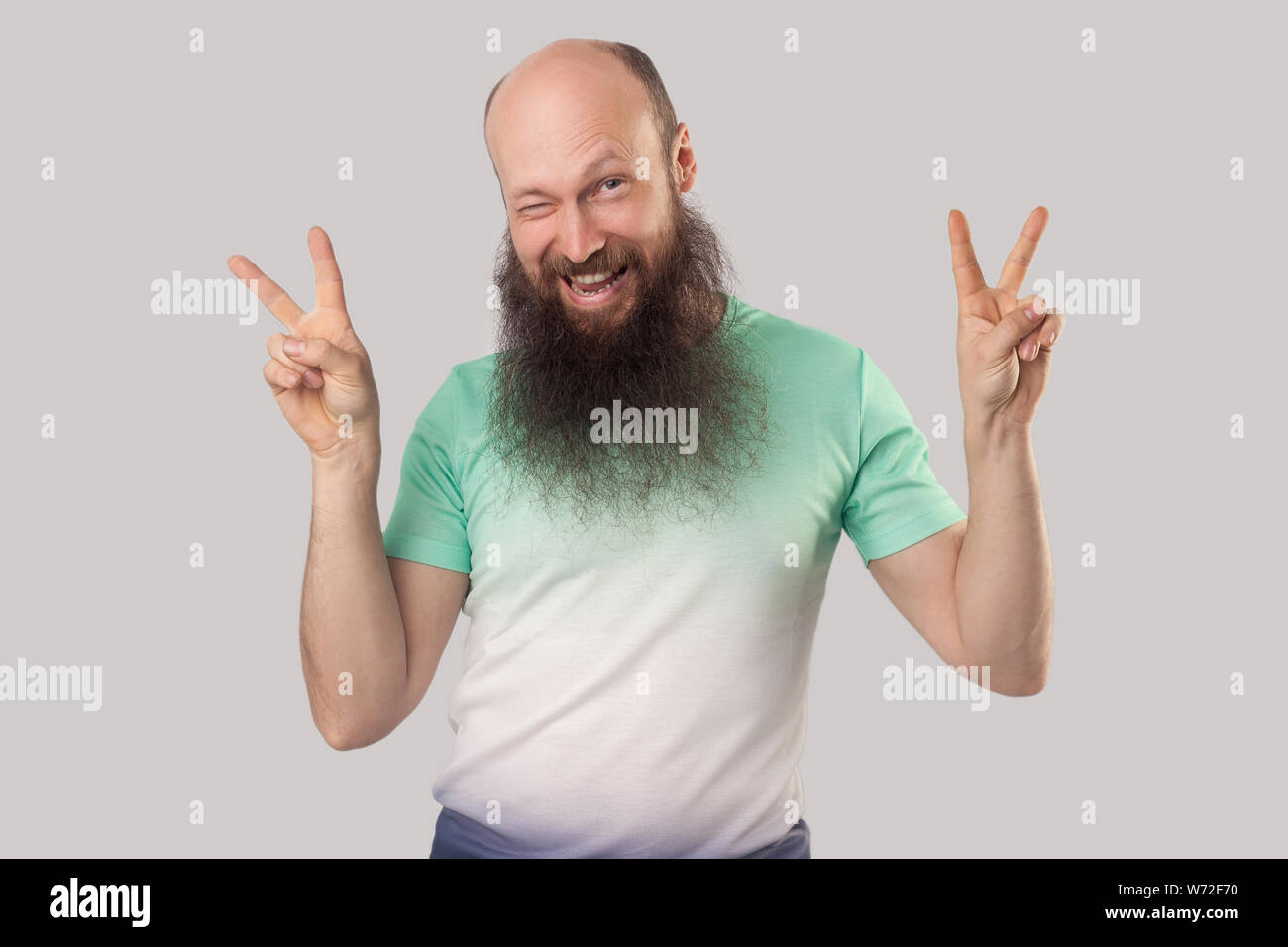 Portrait d'âge moyen drôle homme chauve avec longue barbe en vert clair t-shirt debout avec la victoire ou geste de paix et à la recherche et clignant de l'intérieur. s Banque D'Images