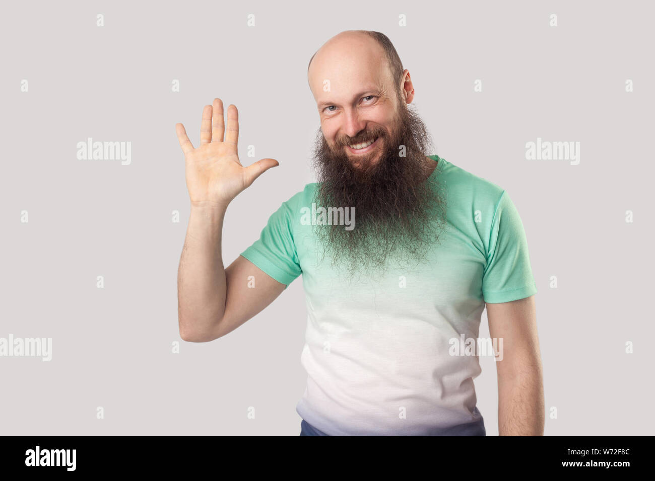Portrait of happy senior homme chauve avec longue barbe en vert clair t-shirt standing looking avec sourire à pleines dents et en agitant sa main et d'accueil. ind Banque D'Images