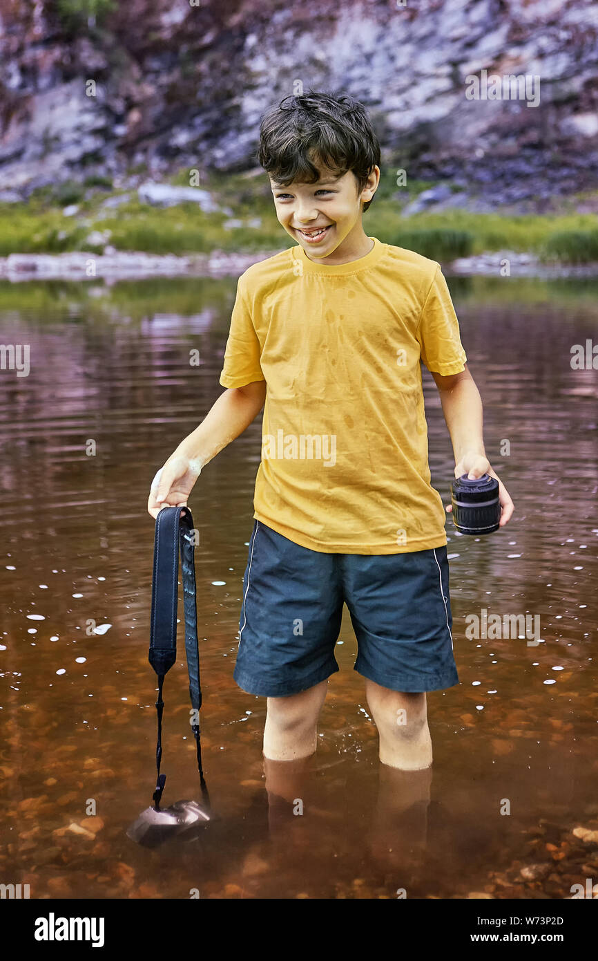 Pretty smiling boy blanc sur 8 ans est debout dans la rivière avec appareil photo numérique en partie dans l'eau, il est gai. Banque D'Images