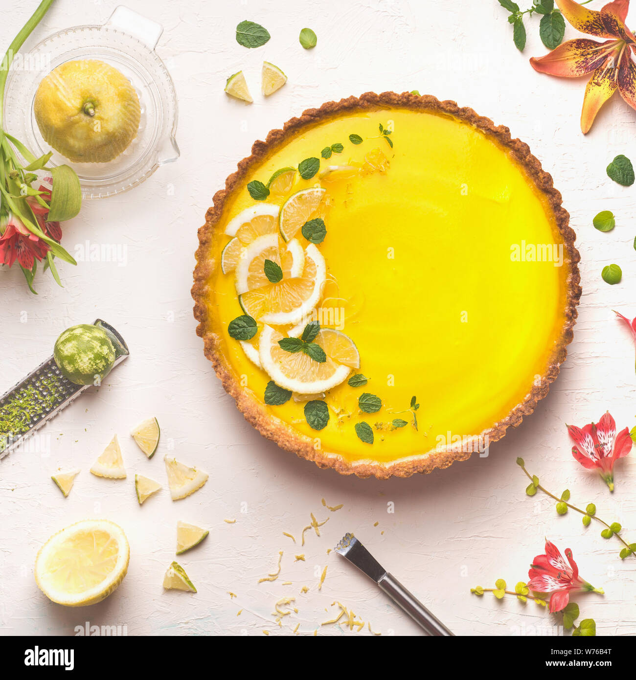 Tarte au citron jaune surmonté de tranches de lime et de citron sur fond de table blanc avec ingrédients agrumes et fleurs, vue d'en haut. Cuisine française Banque D'Images