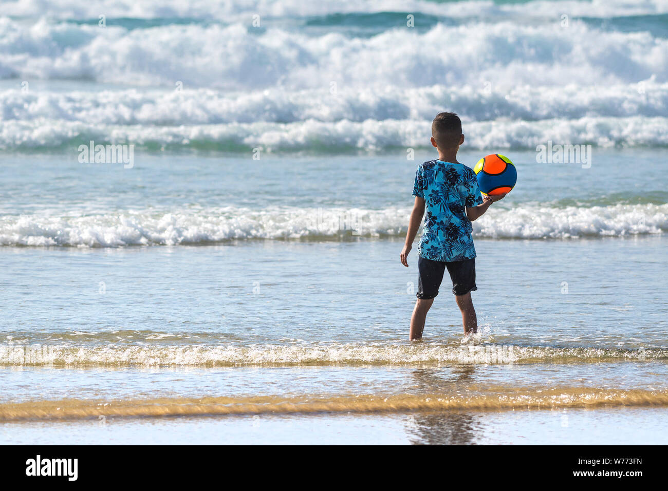 Un jeune garçon debout dans la mer tenant une boule colorée à la plage de Fistral à Newquay en Cornouailles. Banque D'Images