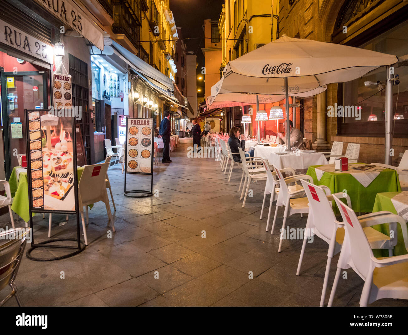 Une rue bordée de cafés et restaurants de nuit à Seviile Espagne avec des gens assis à des tables dehors, sur le trottoir et les serveurs en attente de cust Banque D'Images