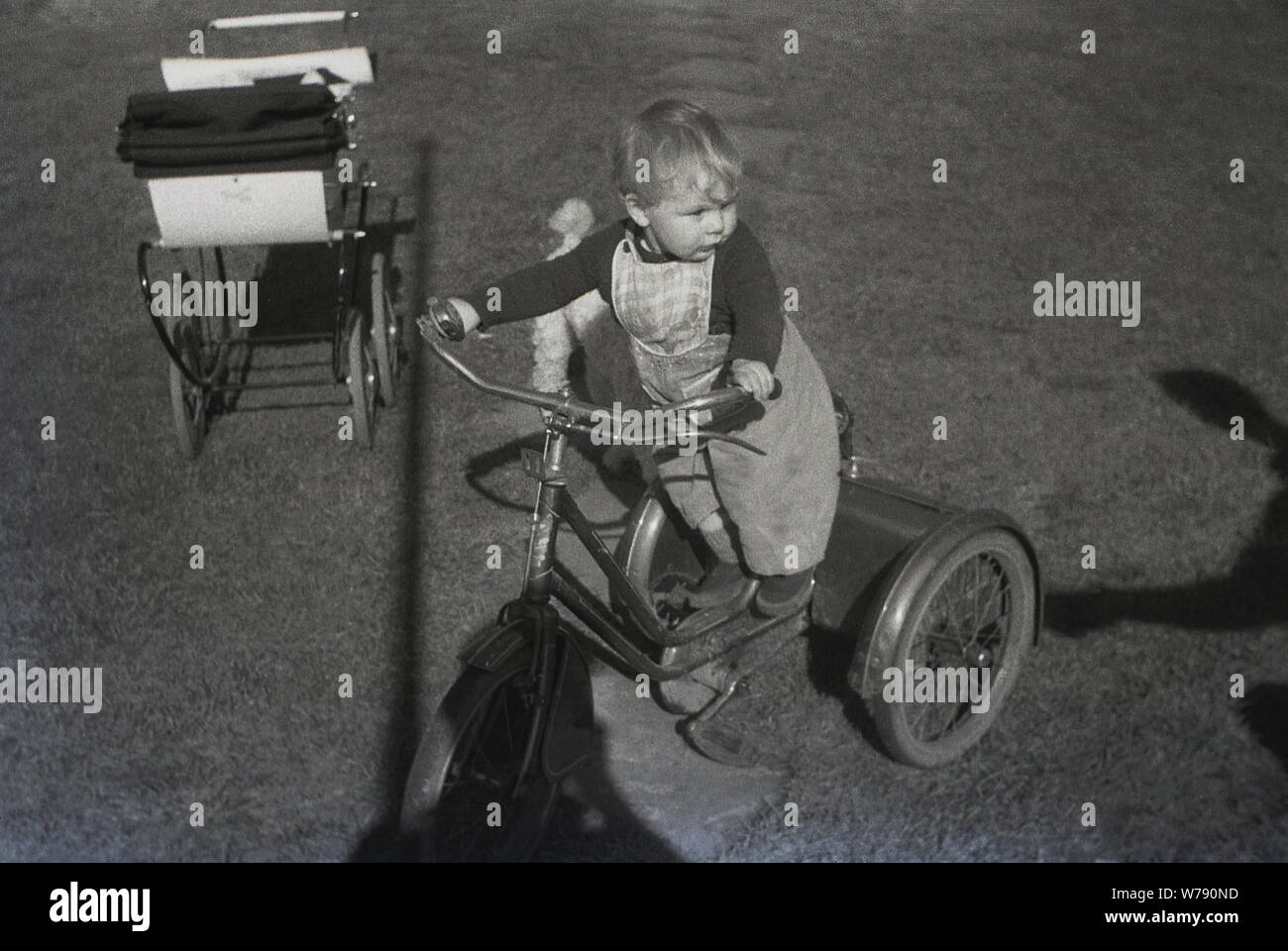 Années 1950, historique, du nourrisson enfant debout sur un tricycle en métal de l'époque à l'extérieur dans un jardin avec coach-construit à côté de la PRAM, England, UK. Banque D'Images