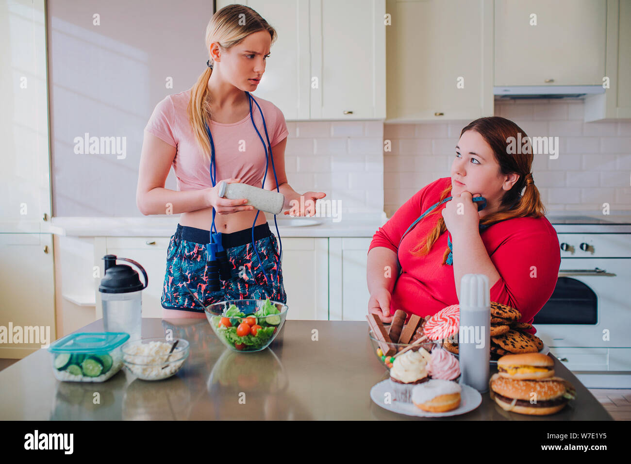 Les jeunes femmes en surpoids et slim regarder les uns les autres dans la cuisine. Modèle mince attente bol avec salade en mains. Grande taille femme s'asseoir et regarder bien-bilt mod Banque D'Images