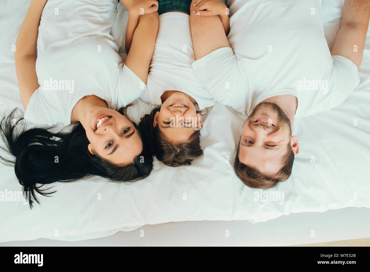 Famille heureuse allongé sur un lit blanc nouveau hugging et à la recherche à l'appareil photo, vue d'en haut. Mère, père et fille lying on bed at home Banque D'Images