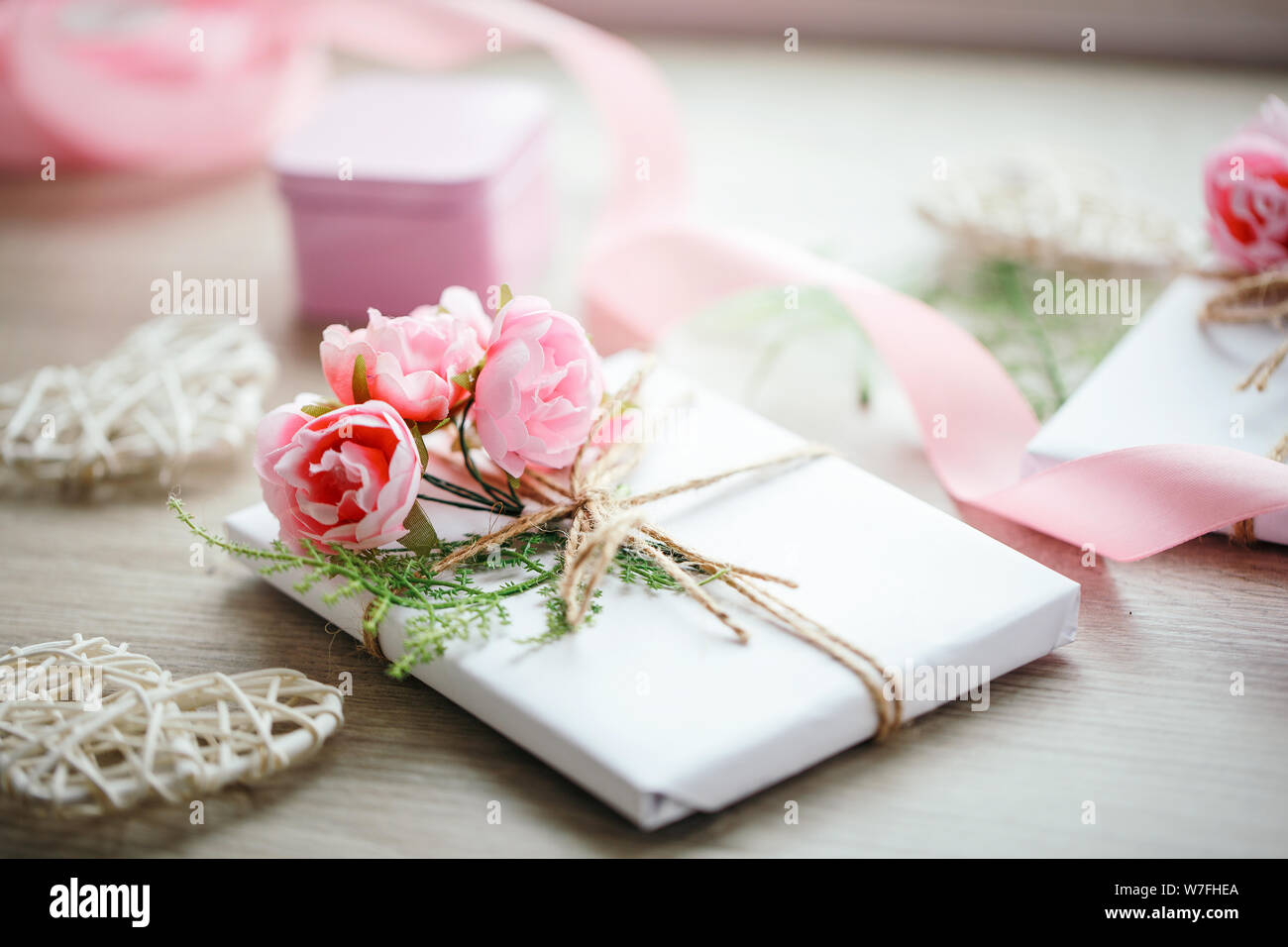 Boîte de cadeau faite à la main enveloppée de papier blanc avec branche de roses. Ruban rose satiné et coeur en bois sur le fond. Banque D'Images