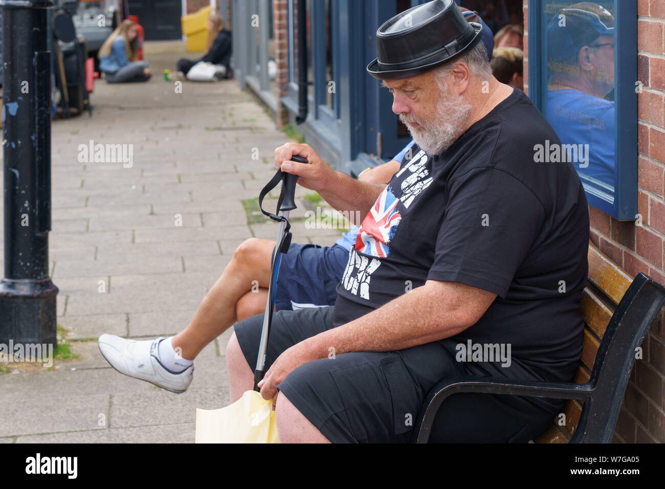 Homme âgé avec une barbe grise assis sur un siège portant un t-shirt punk rock noir et un chapeau en plastique, York, North Yorkshire, Angleterre, Royaume-Uni. Banque D'Images