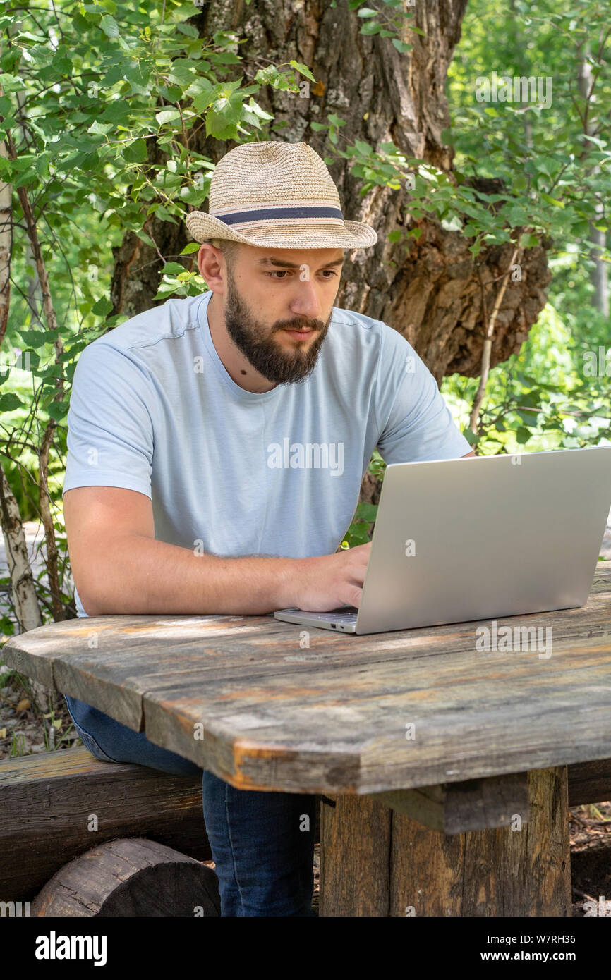 Jeune, barbu dans un chapeau s'assied sur un banc dans l'été dans la rue, à l'aide d'un ordinateur portable, regarder l'écran, un homme travaillant, étudiant ou le surf Banque D'Images