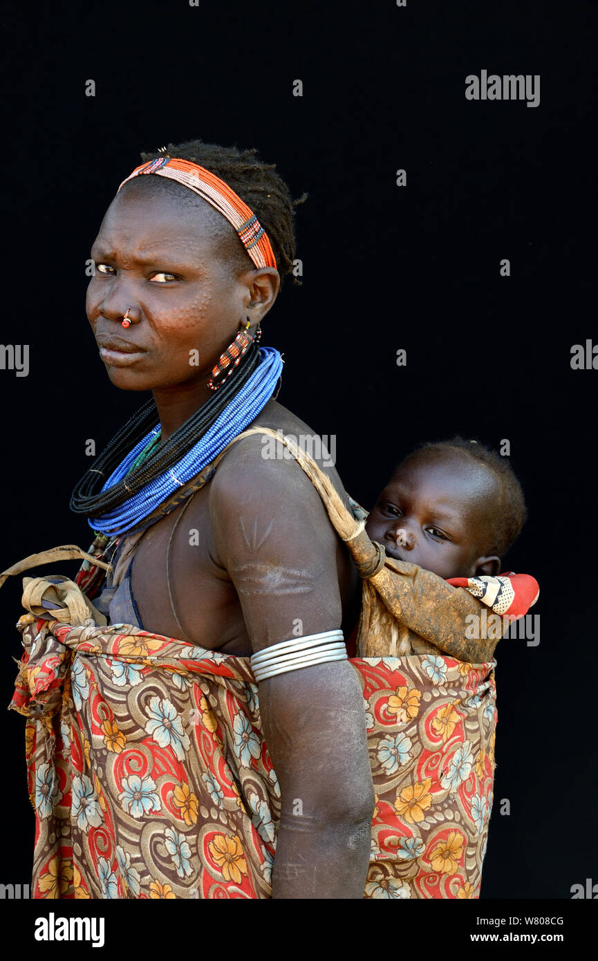 Femme de la tribu Toposas la peau avec scarifications sur le visage et les  bras, et portant des bonnets traditionnels et bijoux, portant son bébé sur  son dos, vallée de l'Omo, en