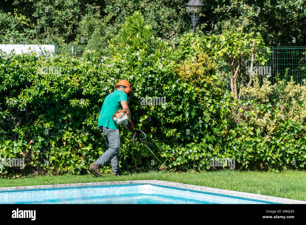 Un homme équipé d'une débroussailleuse travaille sur l'avant cour avec piscine. Concept d'entretien de jardin Banque D'Images