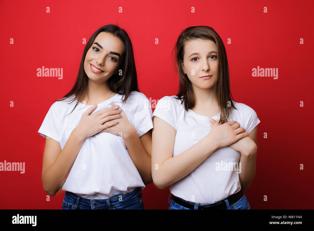 Deux beaux portrait jeune fille vêtue d'une chemise blanche à la recherche en appareil photo avec un visage émotionnel avec les mains de leur cœur contre un mur rouge. Banque D'Images