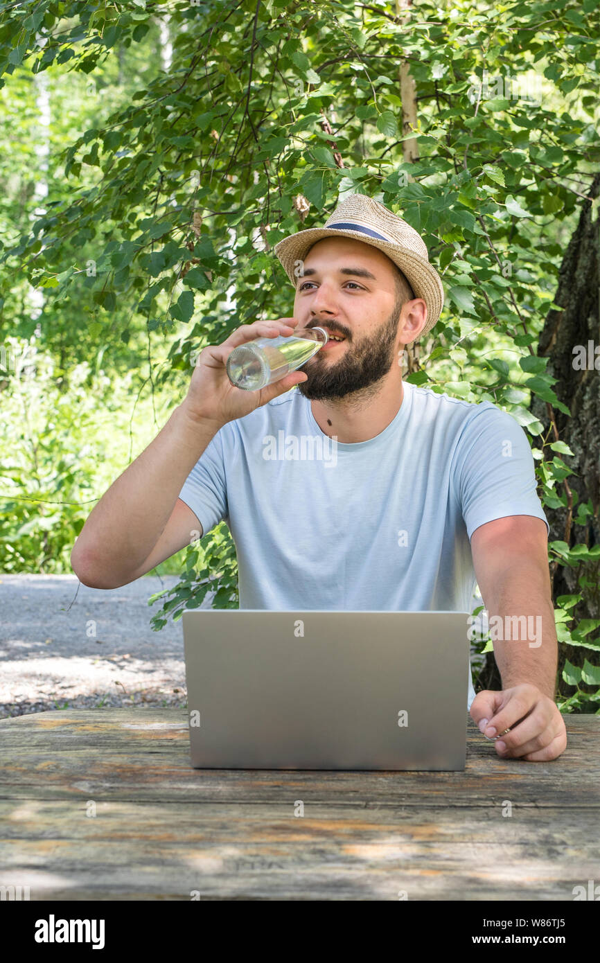 La chaleur dans la rue, soif. Un jeune homme séduisant, avec une barbe d'apparence caucasienne des boissons de l'eau propre à partir d'une bouteille. Assis dans le parc tout en un Banque D'Images