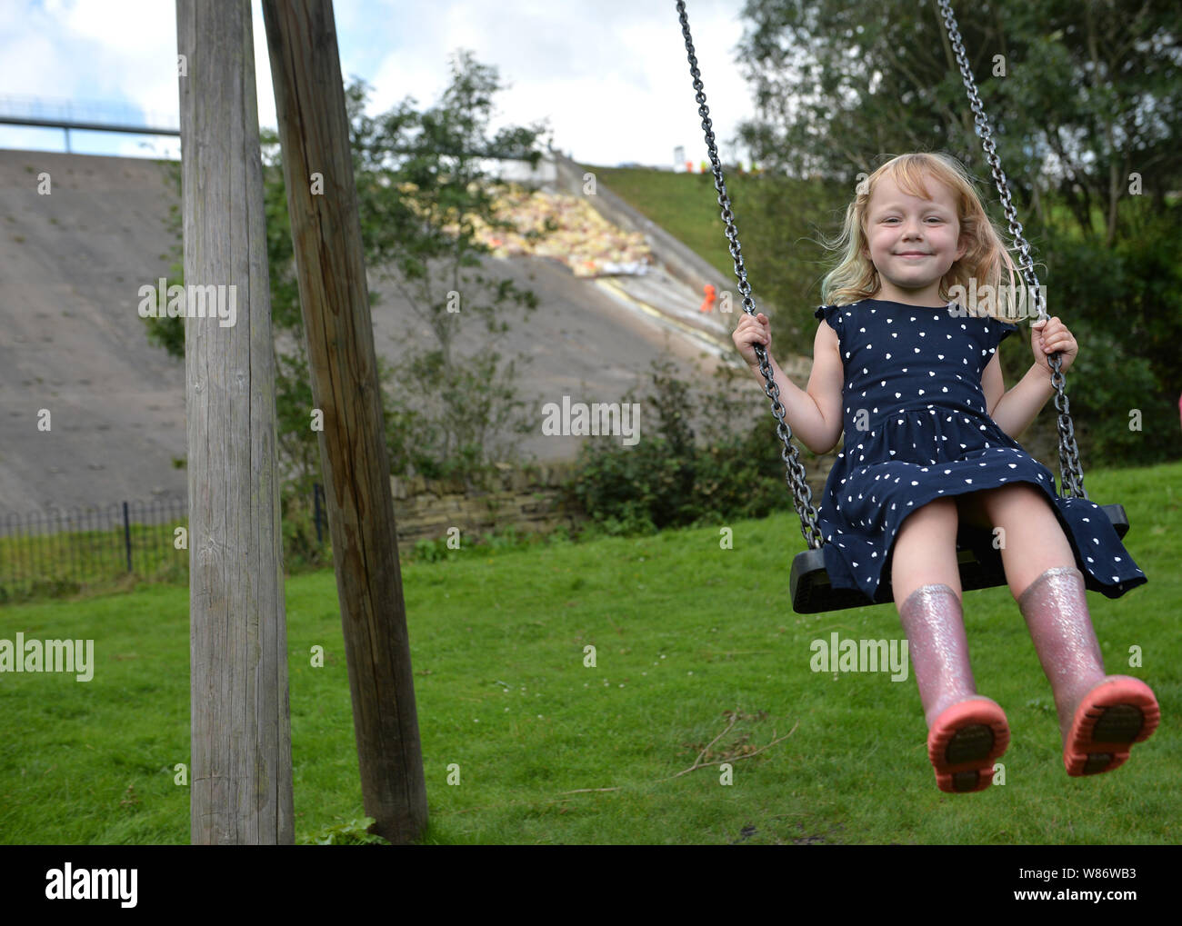 Étant donné la permission des parents, Lala Holden, 5, joue sur une balançoire, tandis que dans le travail de fond continue de soutenir le réservoir du barrage de Toddbrook près du village de Whaley Bridge, Derbyshire, après qu'il a été endommagé d'une forte pluie. Banque D'Images