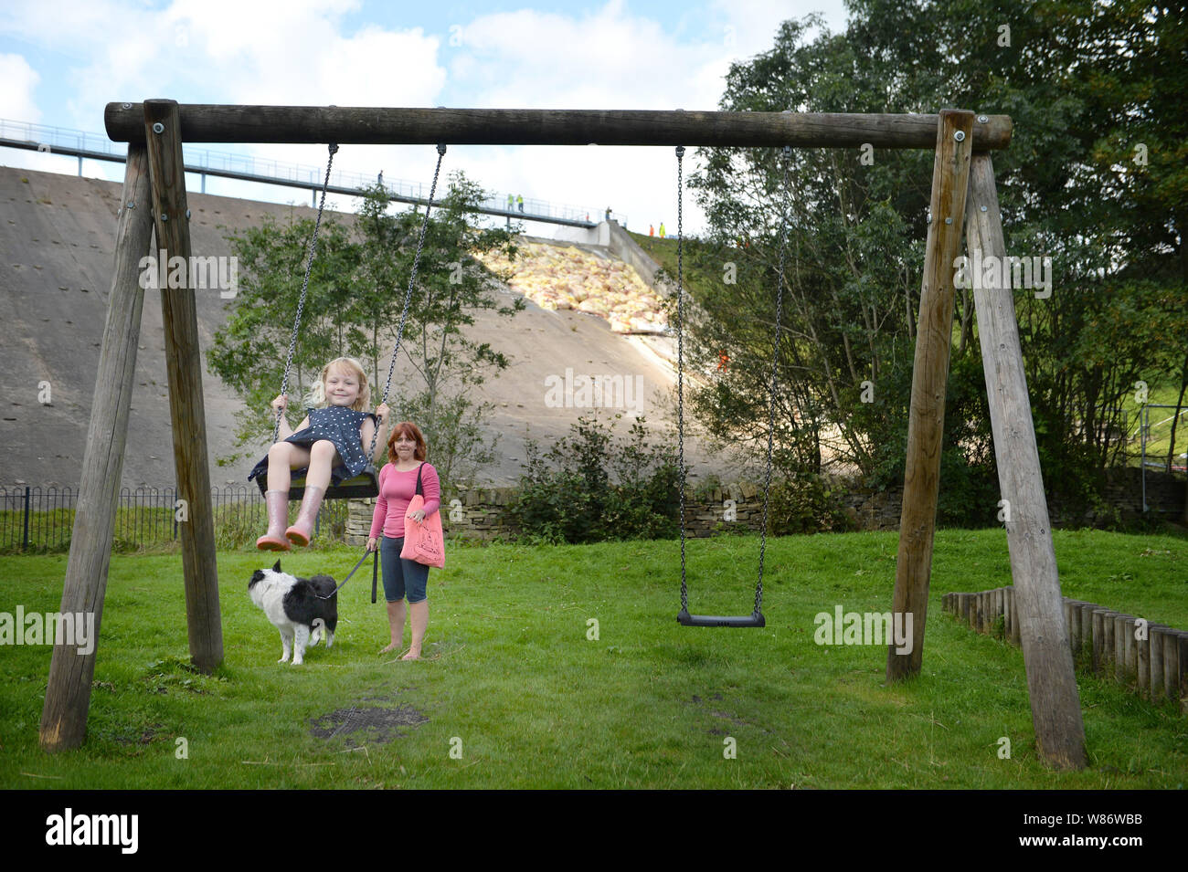 Étant donné la permission des parents, Lala Holden, 5, joue sur une balançoire accompagnée par sa mère, Sally Curley, tandis que dans le travail de fond continue de soutenir le réservoir du barrage de Toddbrook près du village de Whaley Bridge, Derbyshire, après qu'il a été endommagé d'une forte pluie. Banque D'Images