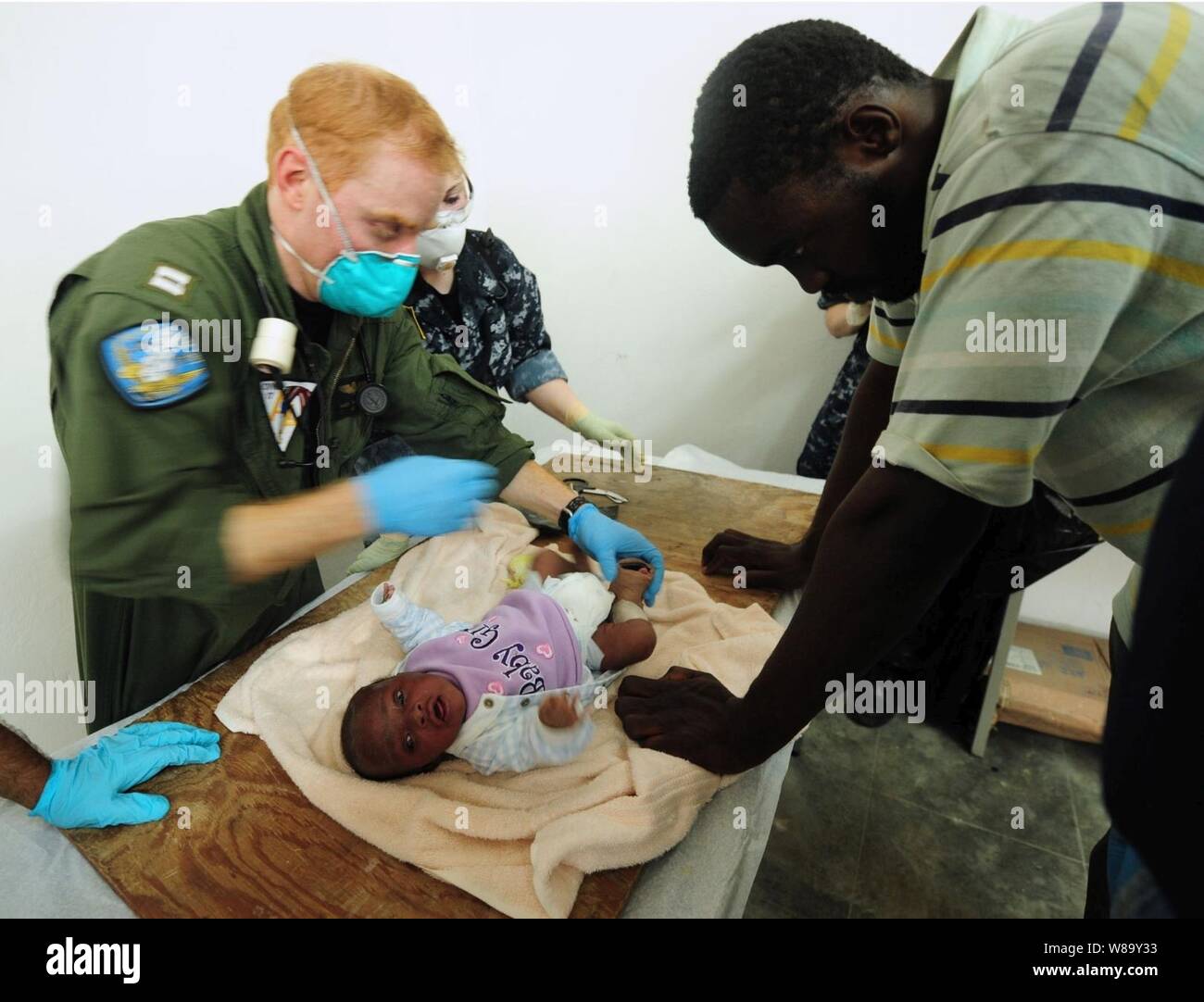 Les marins de la Marine américaine affecté à le porte-avions USS Carl Vinson (CVN 70) soigner un bébé blessé par un tremblement de terre comme le childís montres père à Port-au-Prince, Haïti, le 17 janvier 2010. Le navire et le transporteur Air Wing 17 mènent des opérations de secours en cas de catastrophe humanitaire et dans le cadre de l'opération réponse unifiée après un séisme de magnitude 7,0 a causé de graves dommages le 12 janvier 2010. Banque D'Images