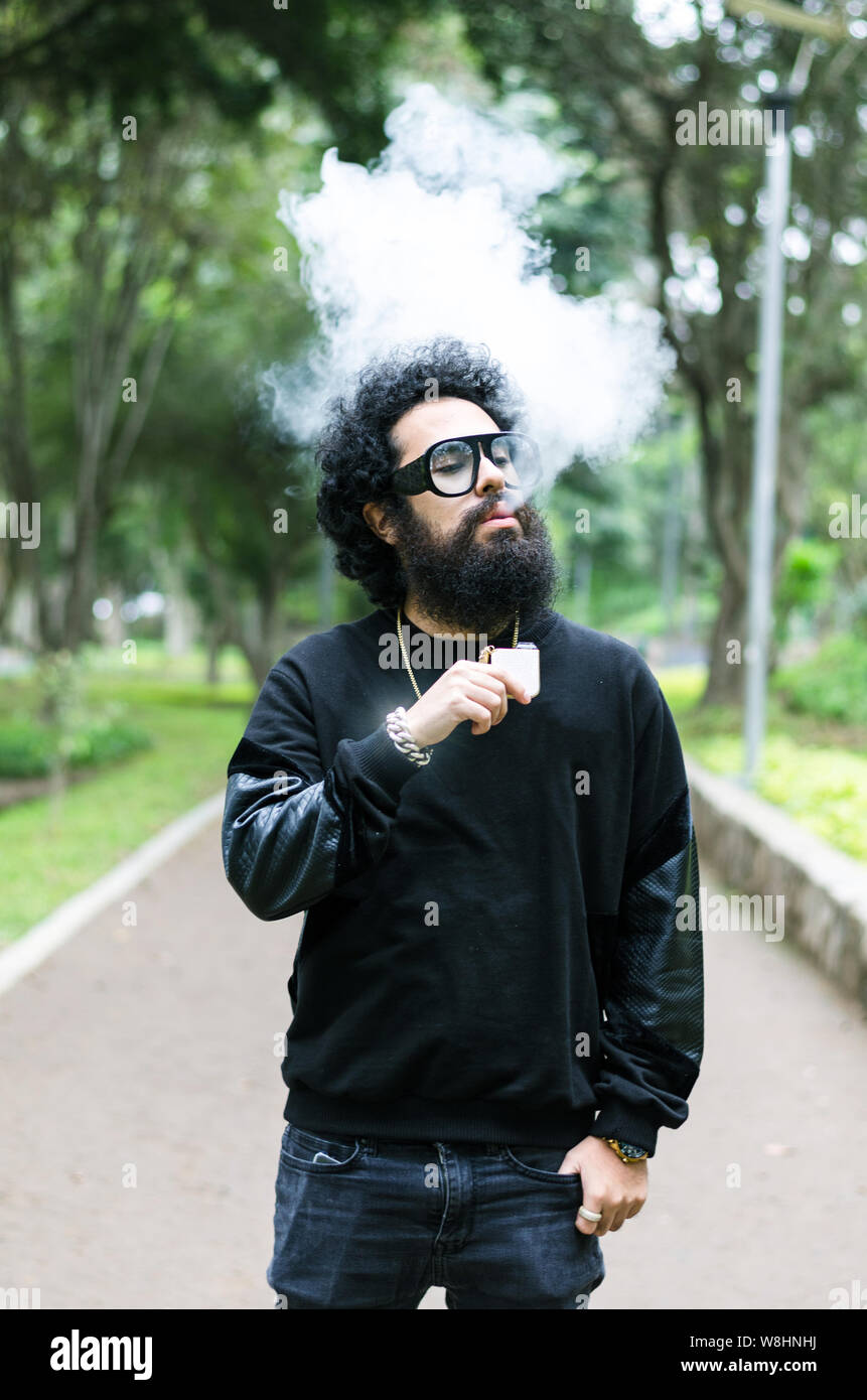 Vape. Jeune homme brutal avec grande barbe et lunettes  la mode coupe de  cheveux dans un tabac cigarette lectronique dans le parc de la ville.  Nuage de vapeur. Style de vie
