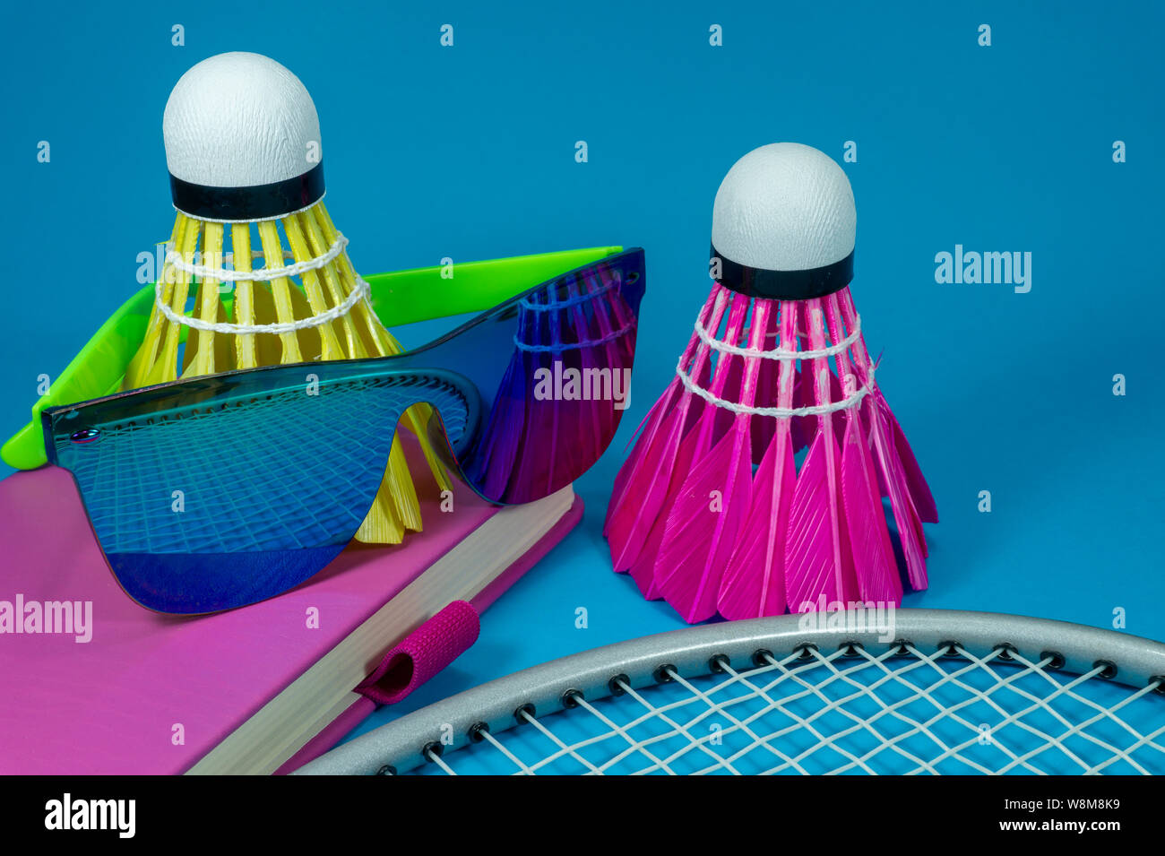 Volants de badminton raquette colorés avec des lunettes de soleil et se reposer sur un livre rose de couleur vive ou de journal sur un fond bleu dans un close up encore Banque D'Images
