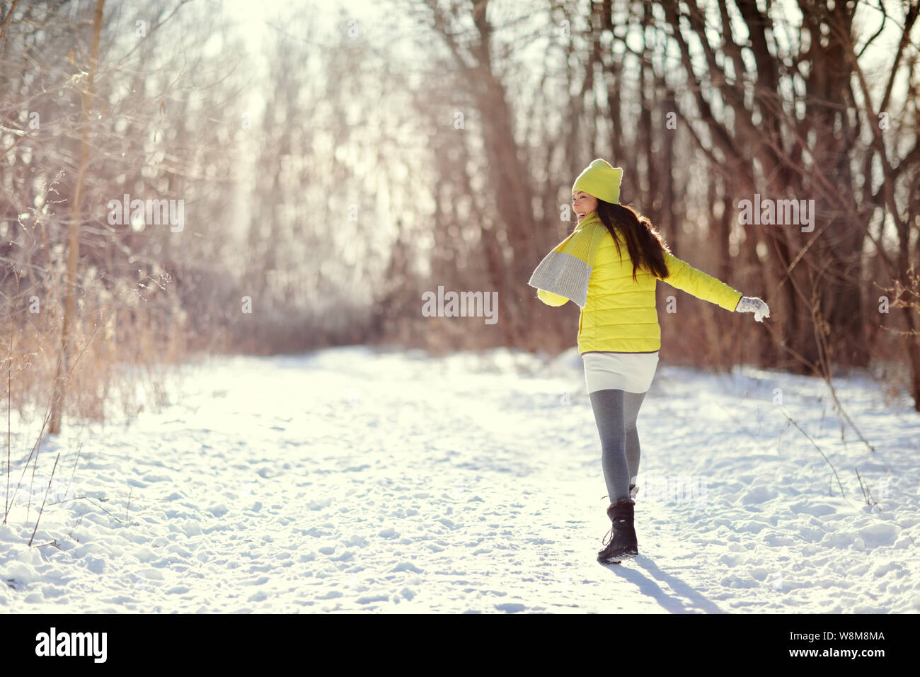 Heureux hiver femme marche dans la neige à l'extérieur de la nature. Jeune femme joyeuse s'amusant sur une activité de plein air à pied sur chemin forestier portant des vêtements veste jaune avec des bottes chaudes, écharpe, chapeau. Banque D'Images