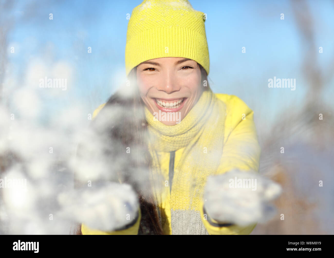 Winter fun girl throwing snow ludique au portrait de l'appareil photo. Asian woman face closeup avec beanie Knit hat jaune et gants blancs jouant avec des flocons de neige. Boule de lutte contre la neige hiver heureux concept. Banque D'Images