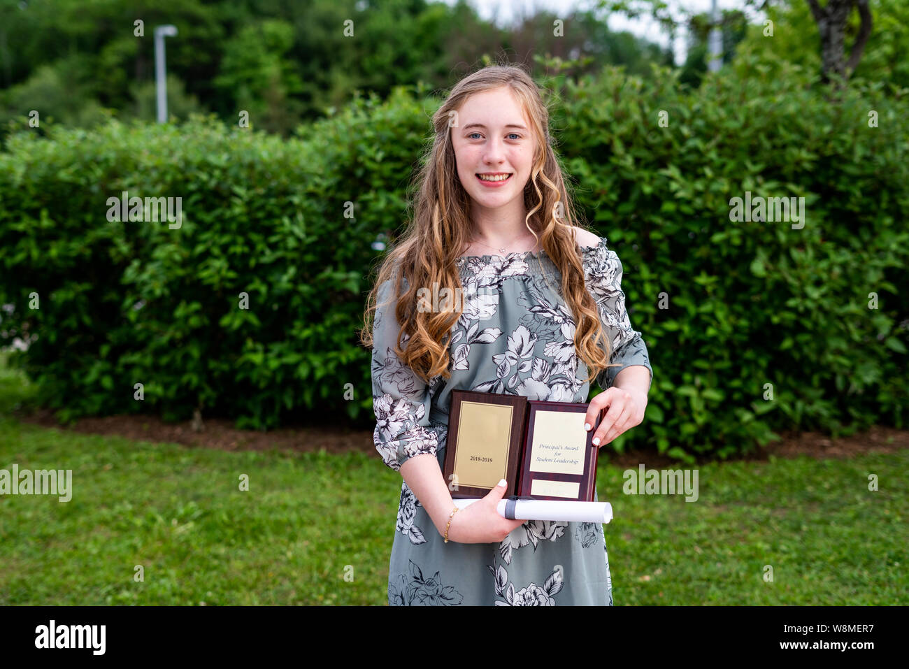 Young teen girl/Middle school student standing in front of a bush/jardin après sa remise des diplômes tout en maintenant un diplôme et prix universitaires. Banque D'Images