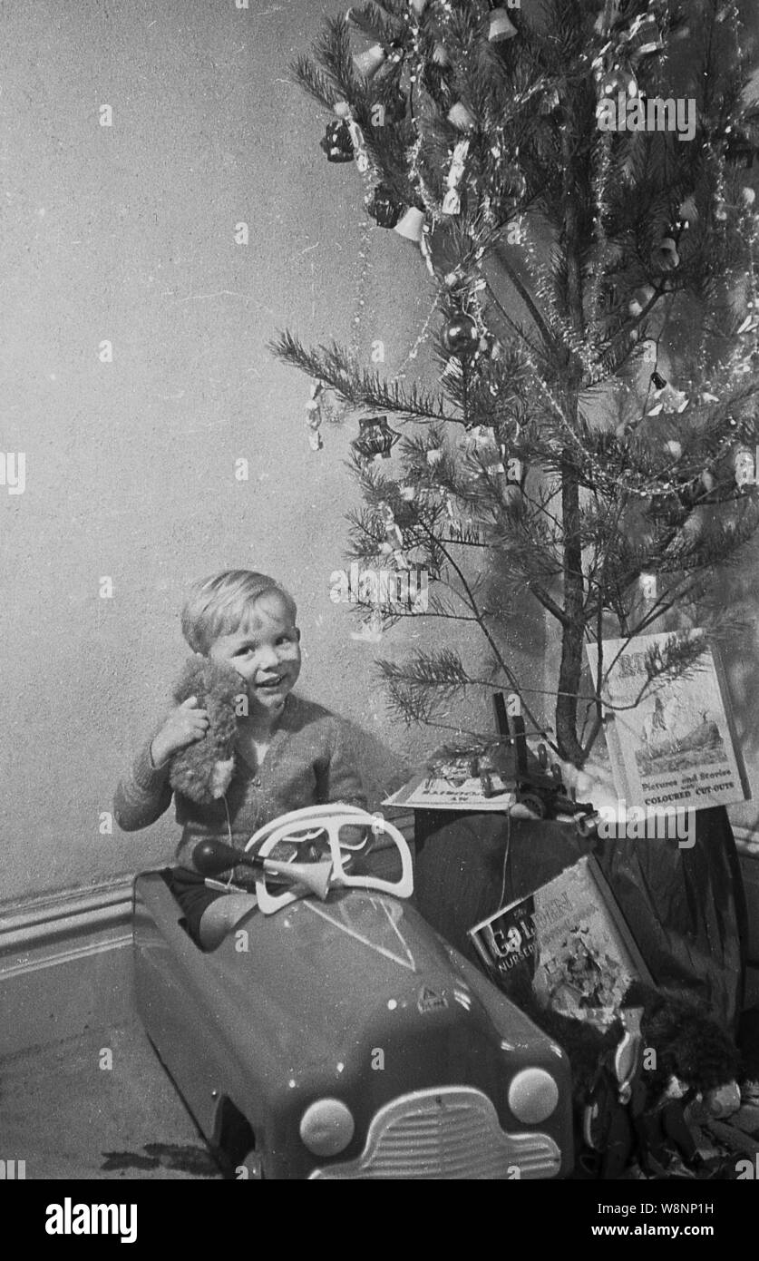 Années 1950, historique, un petit garçon serrant ses équilibrées à porter assis dans sa voiture jouet à conduite à côté d'un arbre de Noël, England, UK. Banque D'Images