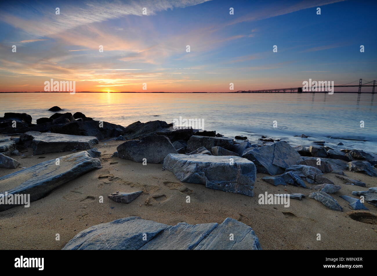 Lever du soleil sur l'horizon au pont de la baie de Chesapeake dans le Maryland. Matin colorés illuminent le ciel des roches au premier plan le long de la côte de sable. Banque D'Images