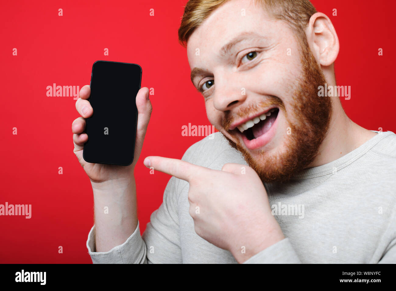 Handsome man with ginger beard pointant sur smartphone Avec écran vide et à la recherche à l'appareil photo avec l'expression visage excité en se tenant sur le re lumineux Banque D'Images