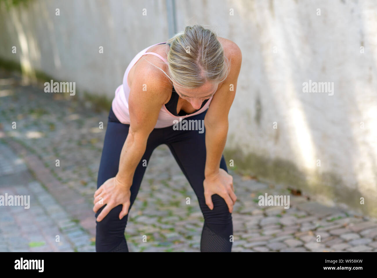 Mettre en place la femme se reposer pendant une séance de pliage vers l'avant avec les mains sur les genoux tandis que dehors marcher dans un chemin ombragé dans un style de vie en plein air actif conc Banque D'Images
