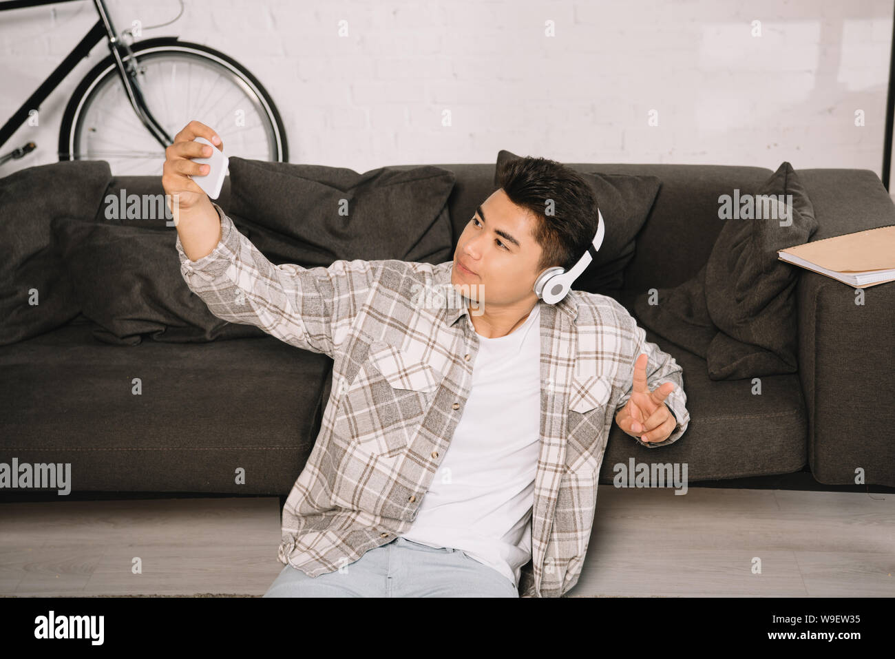 Handsome asian man in headphones sitting on floor et en tenant avec smartphone selfies Banque D'Images