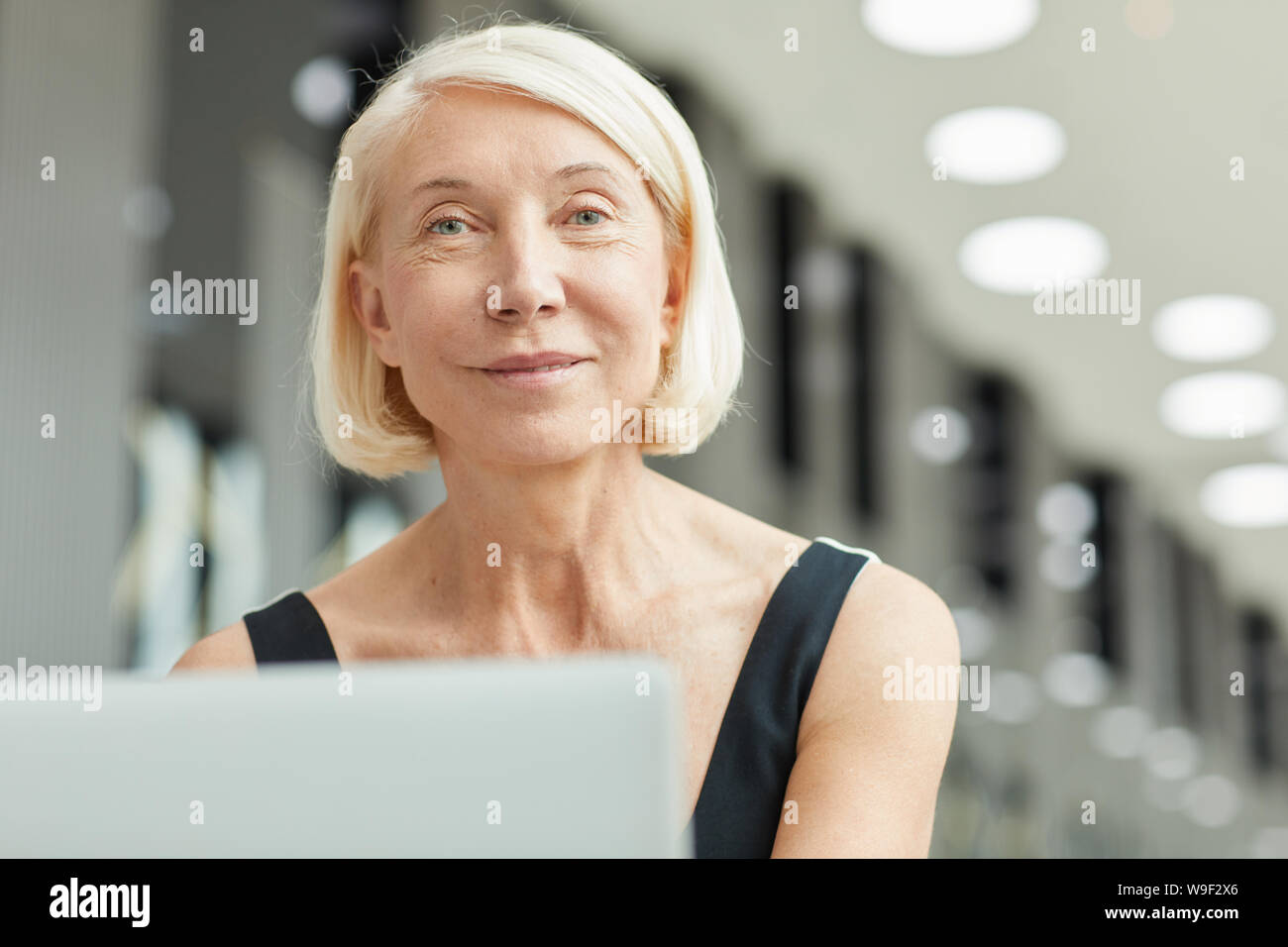 Portrait de femme mature avec de courts cheveux blonds looking at camera while working on laptop computer at office Banque D'Images