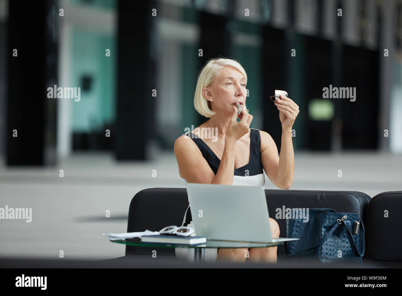 Belle mature businesswoman with blonde cheveux courts assis sur un canapé et appliquer le rouge à lèvres sur ses lèvres avant de rencontrer Banque D'Images