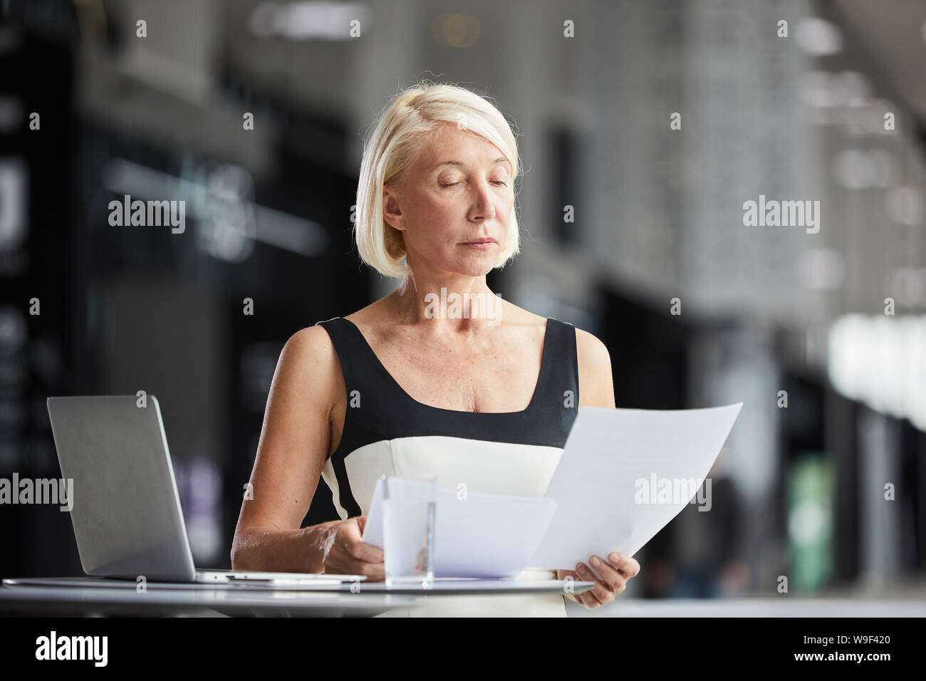 Femme sérieuse avec de courts cheveux blonds assis à la table en face de l'ordinateur portable et de la lecture du contrat d'entreprise Banque D'Images