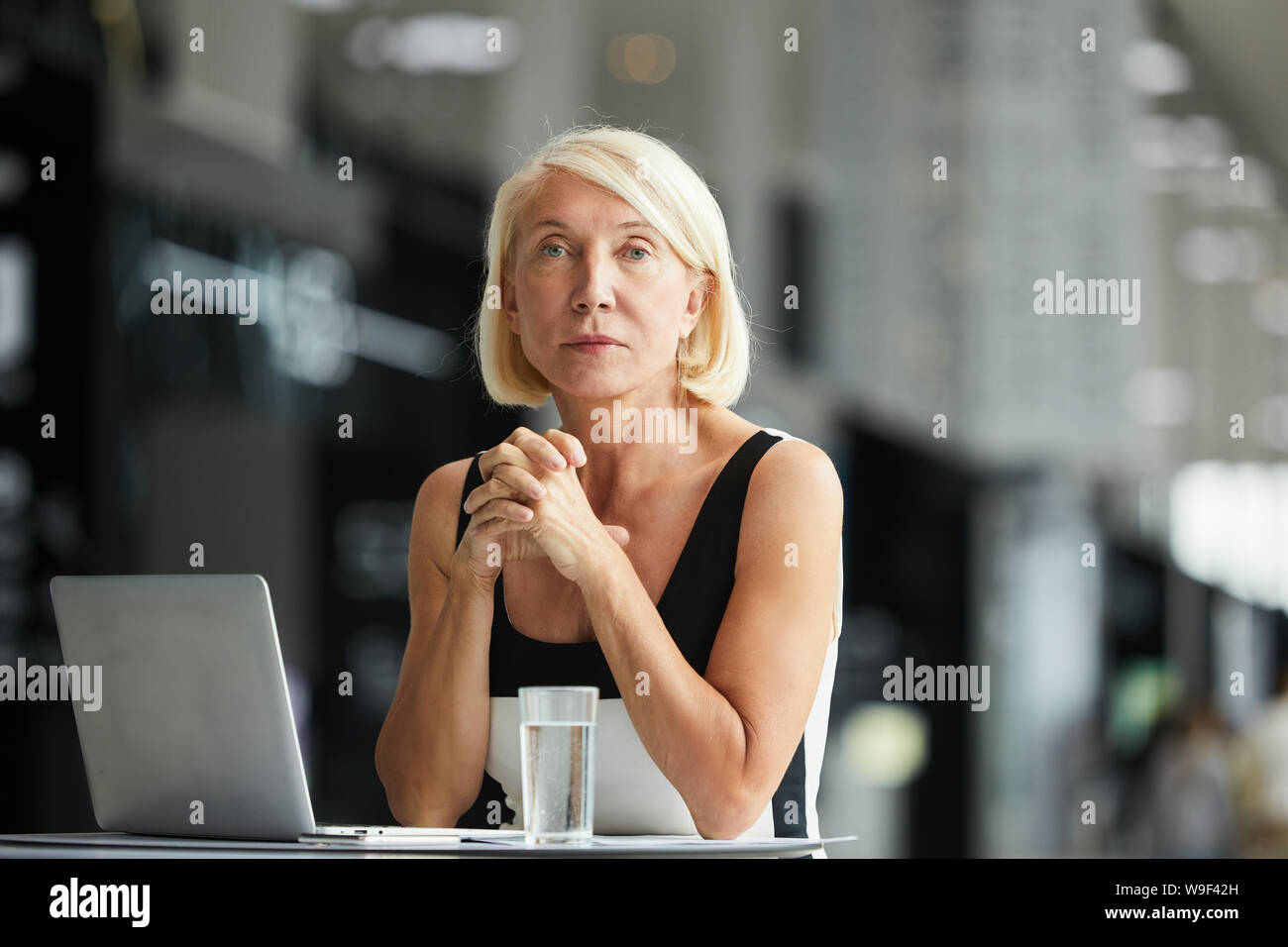 Portrait de femme mature avec de courts cheveux blonds looking at camera, assis à la table avec laptop at office Banque D'Images