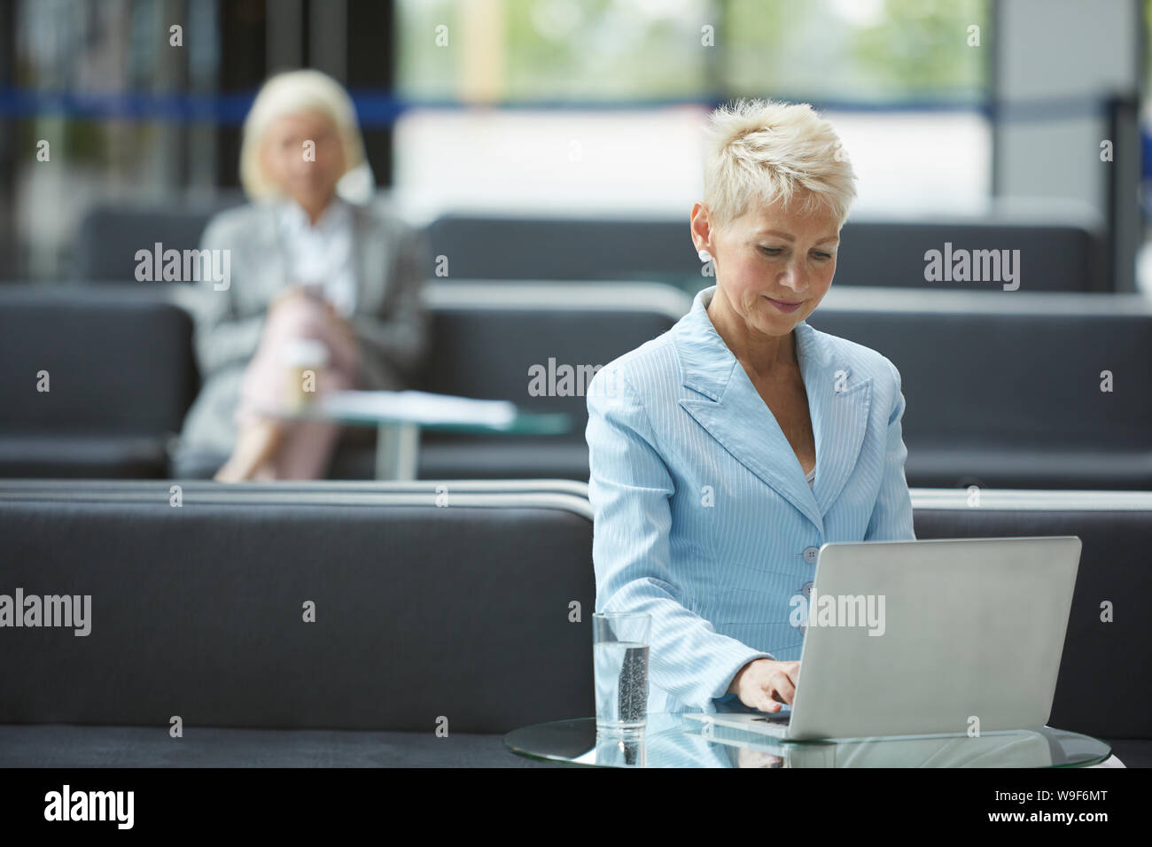 Femme mature avec de courts cheveux blonds assis à la table et de la saisie sur ordinateur portable avec d'autres femme assise à l'arrière-plan à l'office de tourisme Banque D'Images