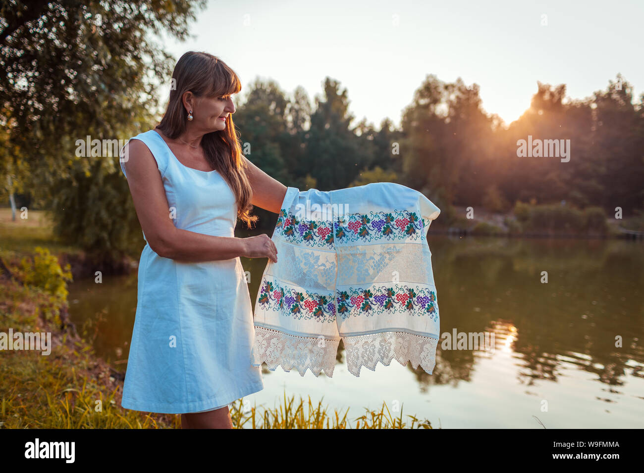 Mature Woman holding serviette avec broderie traditionnelle ukrainienne en face de river in autumn Park au coucher du soleil. Senior woman est fier de sa nationalité Banque D'Images
