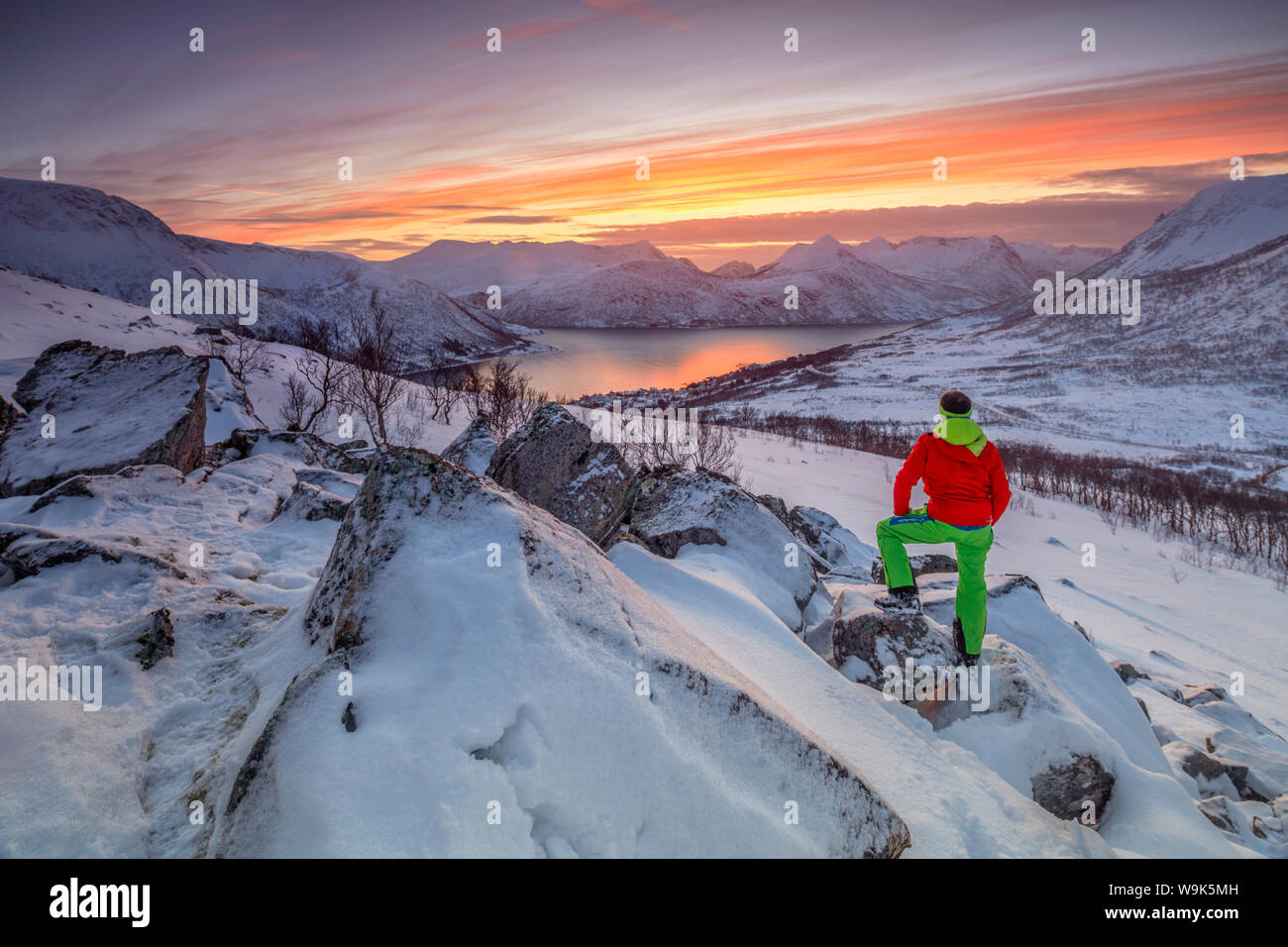 Randonneur admire la mer gelée entouré par la neige encadré par le ciel orange au coucher du soleil, de Torsken, Senja, Troms County, arctique, Norway, Scandinavia, Europe Banque D'Images