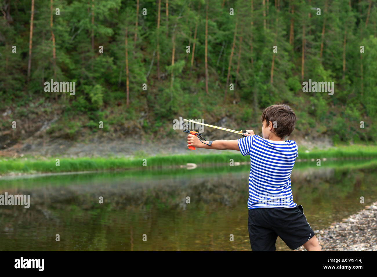 9 ans avec une fronde dans ses mains sur la toile de fond d'un paysage de montagne. Le garçon s'attaque et s'apprête à tourner une fronde. Banque D'Images